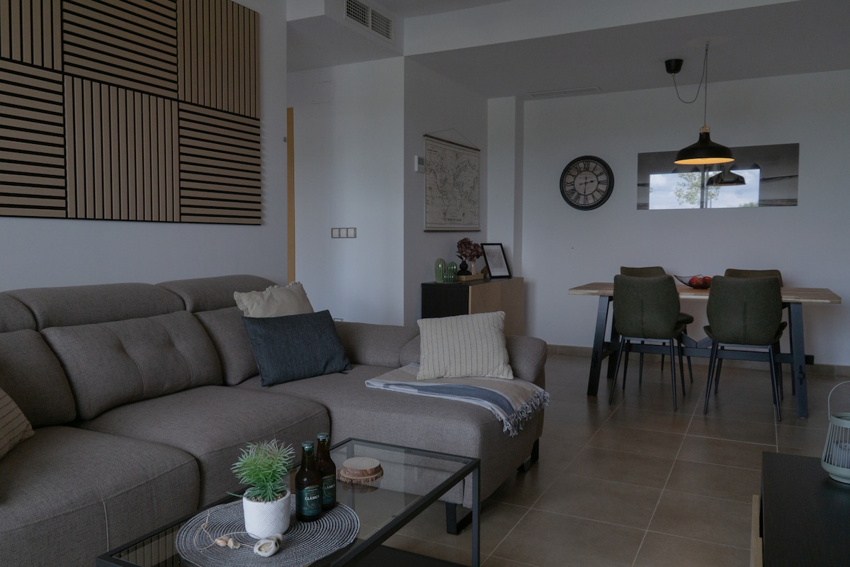 A modern open living area features a comfortable sectional sofa adorned with decorative pillows. A glass coffee table holds small decorative items. In the background, a dining table with four chairs is visible, accompanied by a wall clock and minimalistic art.