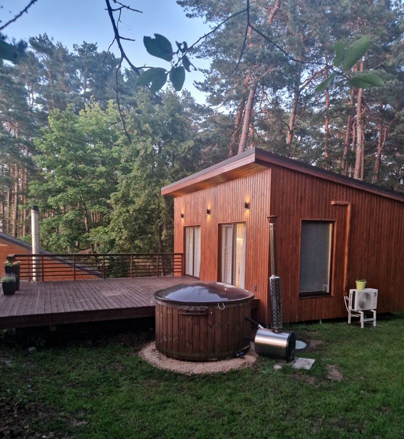 A wooden exterior cabin sits surrounded by tall trees, showcasing large windows. A wooden deck extends from the structure, with a round hot tub positioned nearby. A quiet, natural setting is emphasized, creating a serene atmosphere close to nature.