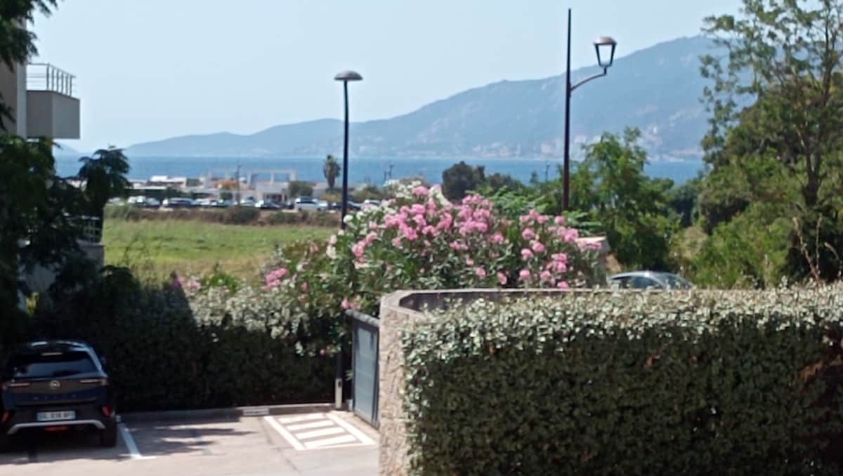 A view framed by lush green foliage features blooming pink flowers, with distant mountains and the sea visible. A small parking area is situated in the foreground, alongside a neatly maintained path, providing a serene backdrop for the surroundings.