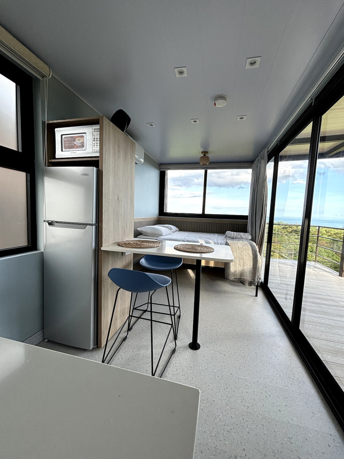 A modern kitchenette area features a compact refrigerator and microwave. A small dining table with two stools is situated near the floor-to-ceiling windows, offering views of the surrounding landscape. A cozy bed is positioned in the corner, adorned with soft linens.