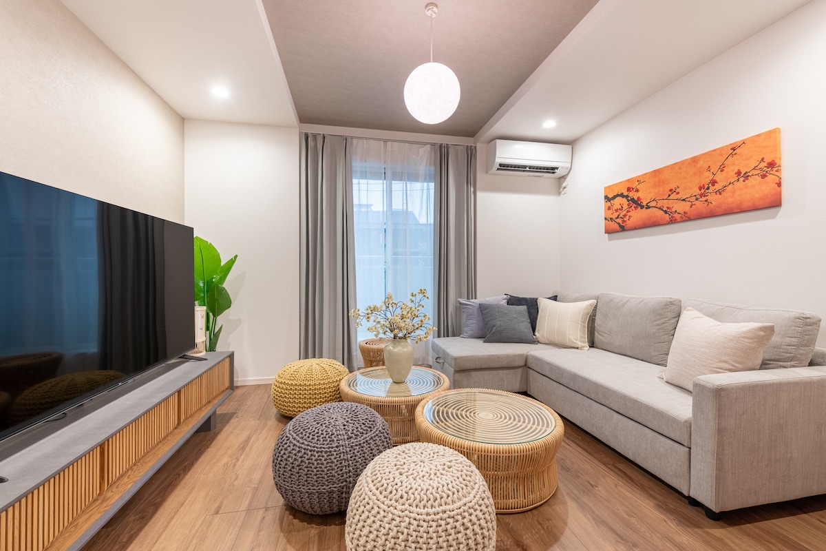 The living room features a sectional sofa accompanied by a mix of poufs in varying textures and colors. A large flat-screen TV is positioned on a wooden console, while a vase of dried flowers sits on a coffee table. Natural light filters through the sheer curtains.