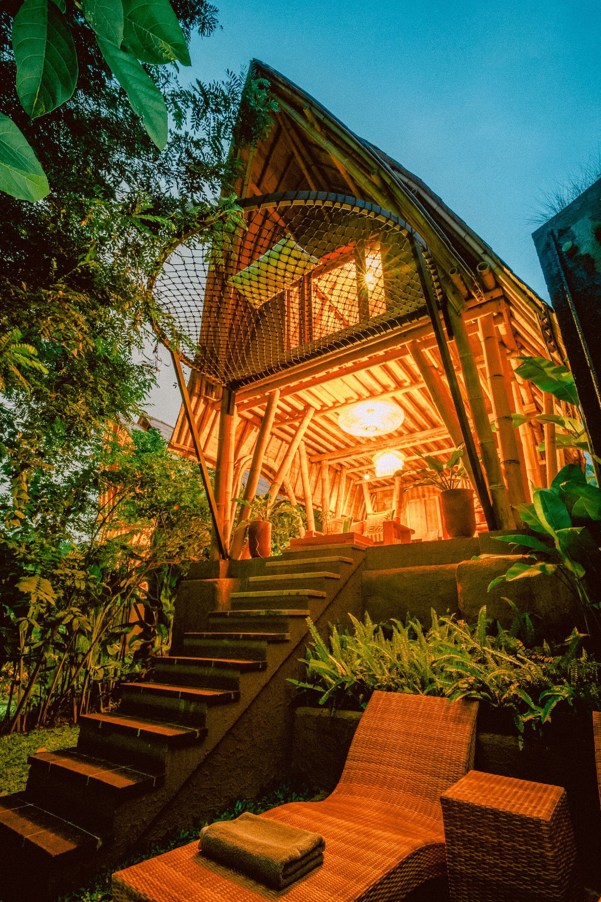 A bamboo house is nestled among lush greenery, illuminated by warm light as evening approaches. Steps lead up to the entrance, complemented by surrounding plants. A comfortable seating area is positioned nearby, inviting relaxation in the natural setting.