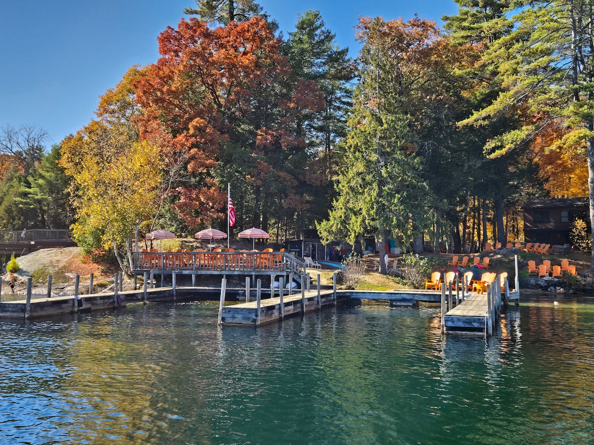 A serene lakeside scene features wooden docks extending into clear water. Colorful autumn foliage surrounds the area, with outdoor seating and umbrellas placed for relaxation. An American flag gently waves near the dock, contributing to the natural beauty of the setting.