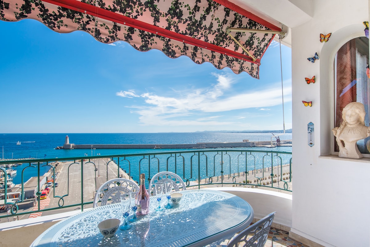 An inviting terrace offers a dining table set with delicate chairs, overlooking a panoramic view of the sea. Bright blue waters stretch to the horizon, with a calm sky above. Decorative elements, including colorful butterflies, add a whimsical touch to the space.