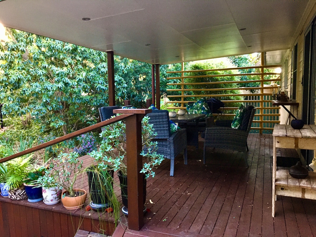 An outdoor covered deck is framed by lush greenery, featuring a dining table surrounded by four woven chairs. Potted plants decorate the area, adding color and life to the space. Natural wood flooring complements the inviting atmosphere of the setting.