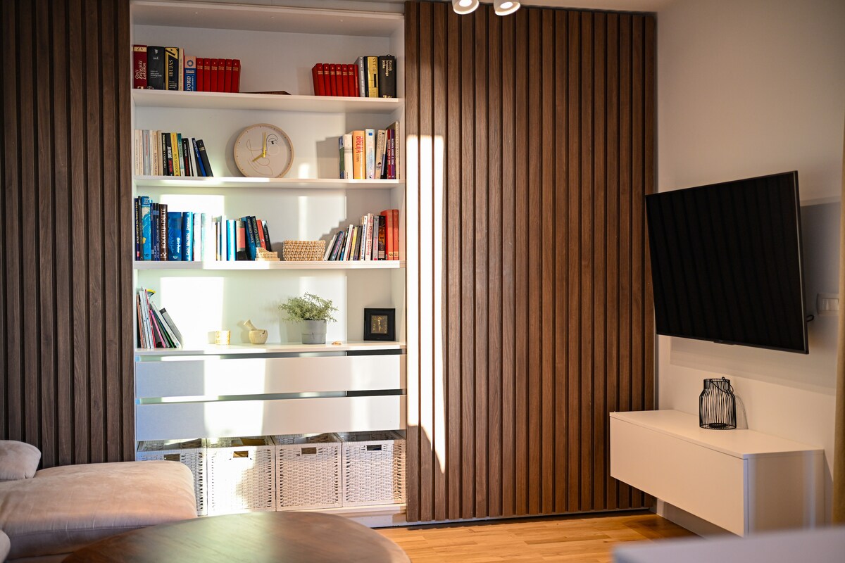 A modern living room is showcased, featuring a wall-mounted television opposite a sleek white shelving unit. The unit is organized with books and decorative items, while a warm wooden accent wall adds texture. Soft lighting enhances the cozy atmosphere.