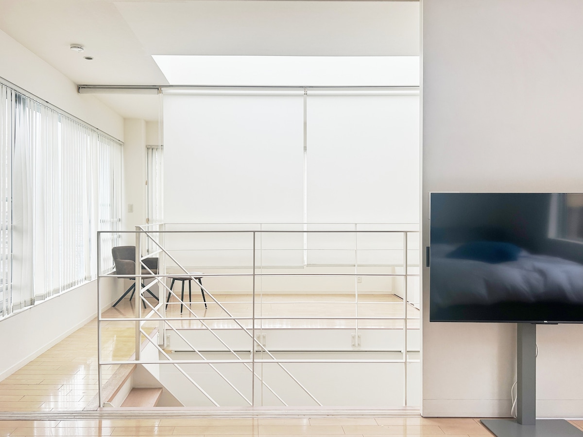 A bright, open space is featured, showcasing natural light filtering through large windows with white blinds. A black chair is visible in the corner, while a television stands on a sleek stand, providing a modern touch to the minimalist design.