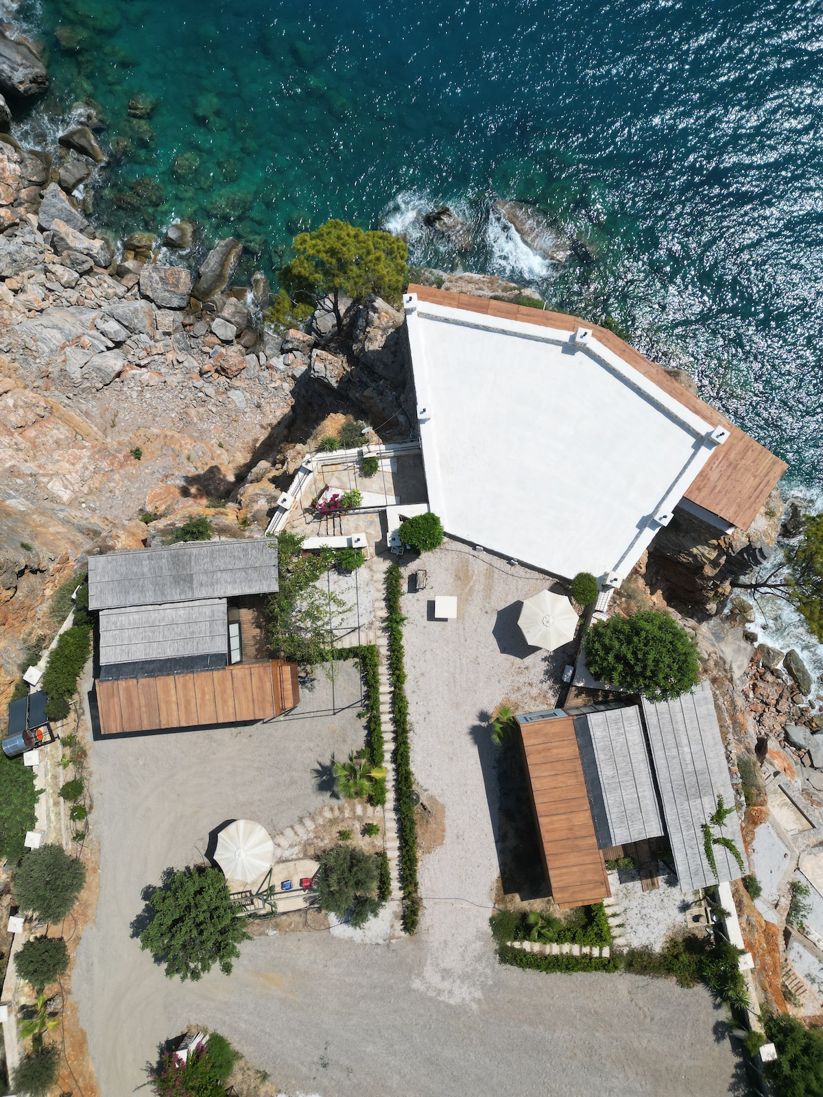 An aerial view captures the unique layout of a coastal retreat, with a hexagonal building situated prominently. Surrounding gardens and pathways are visible, alongside a sandy area with loungers, nestled among natural rock formations and clear blue waters.
