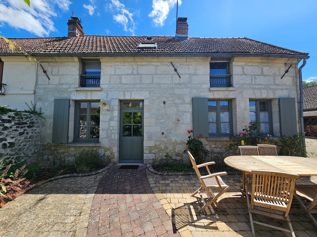 An independent stone house is nestled in a communal courtyard, featuring green shutters and a welcoming front door. A round wooden table with chairs is positioned outside, surrounded by garden areas with flowers and plants. The blue sky above enhances the outdoor setting.