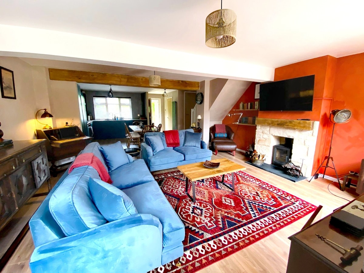 A spacious living area features a large blue sectional sofa arranged around a wooden coffee table. A colorful rug lies beneath, and a stone fireplace is positioned against a contrasting orange wall. Light streaming from windows highlights the cozy atmosphere of the room.