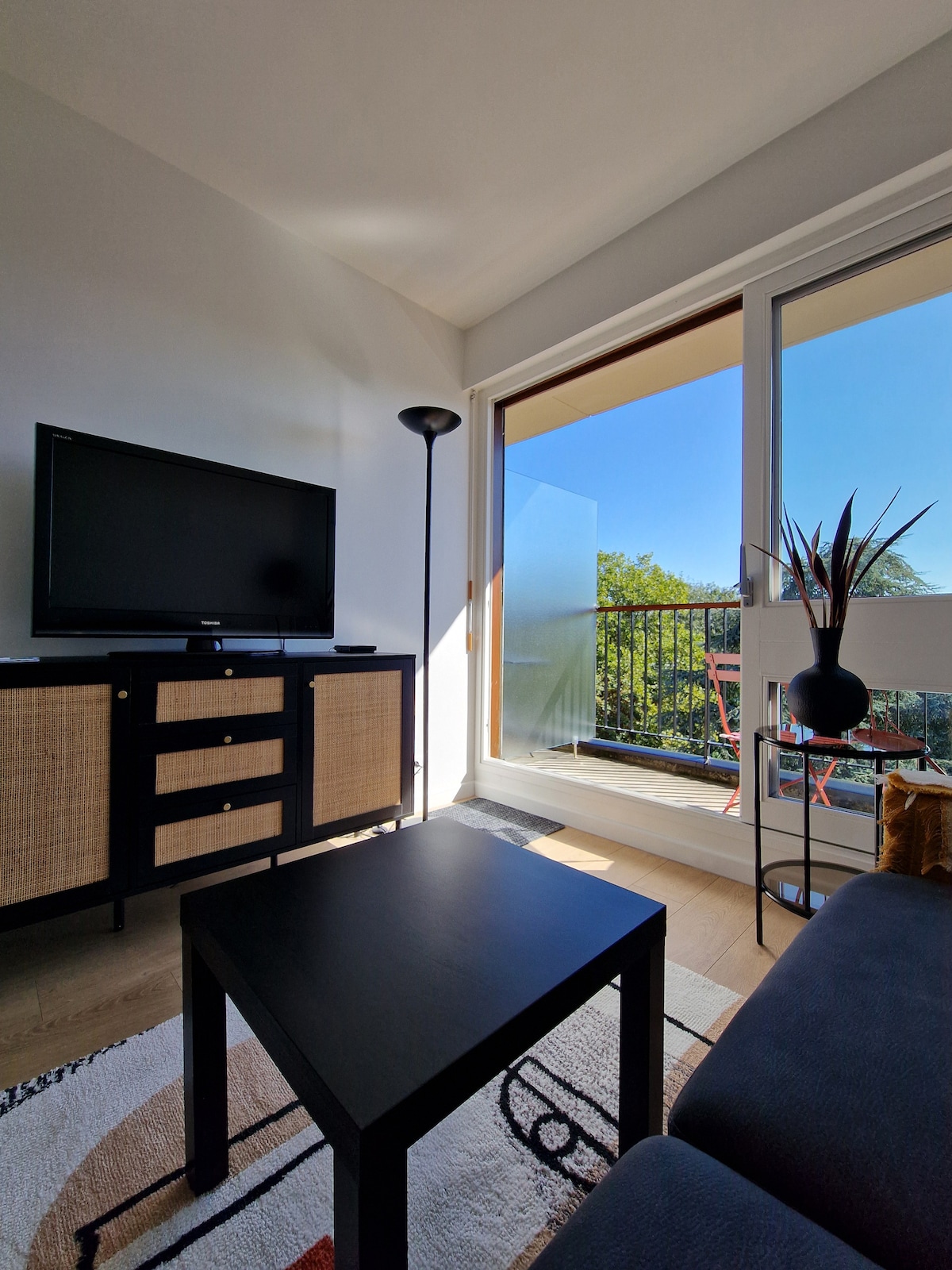 A living area is shown with a black television atop a cabinet featuring wicker details. A small black table is positioned in front of a comfortable sofa. Glass doors lead to a balcony with greenery visible beyond.