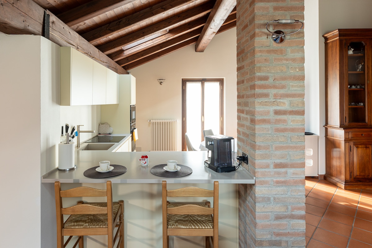 An open kitchen area features wooden beams overhead, creating a rustic feel. A breakfast bar with two wooden chairs is positioned in front of a counter. Appliances such as a coffee maker and microwave are visible, alongside a window allowing natural light to enter the space.
