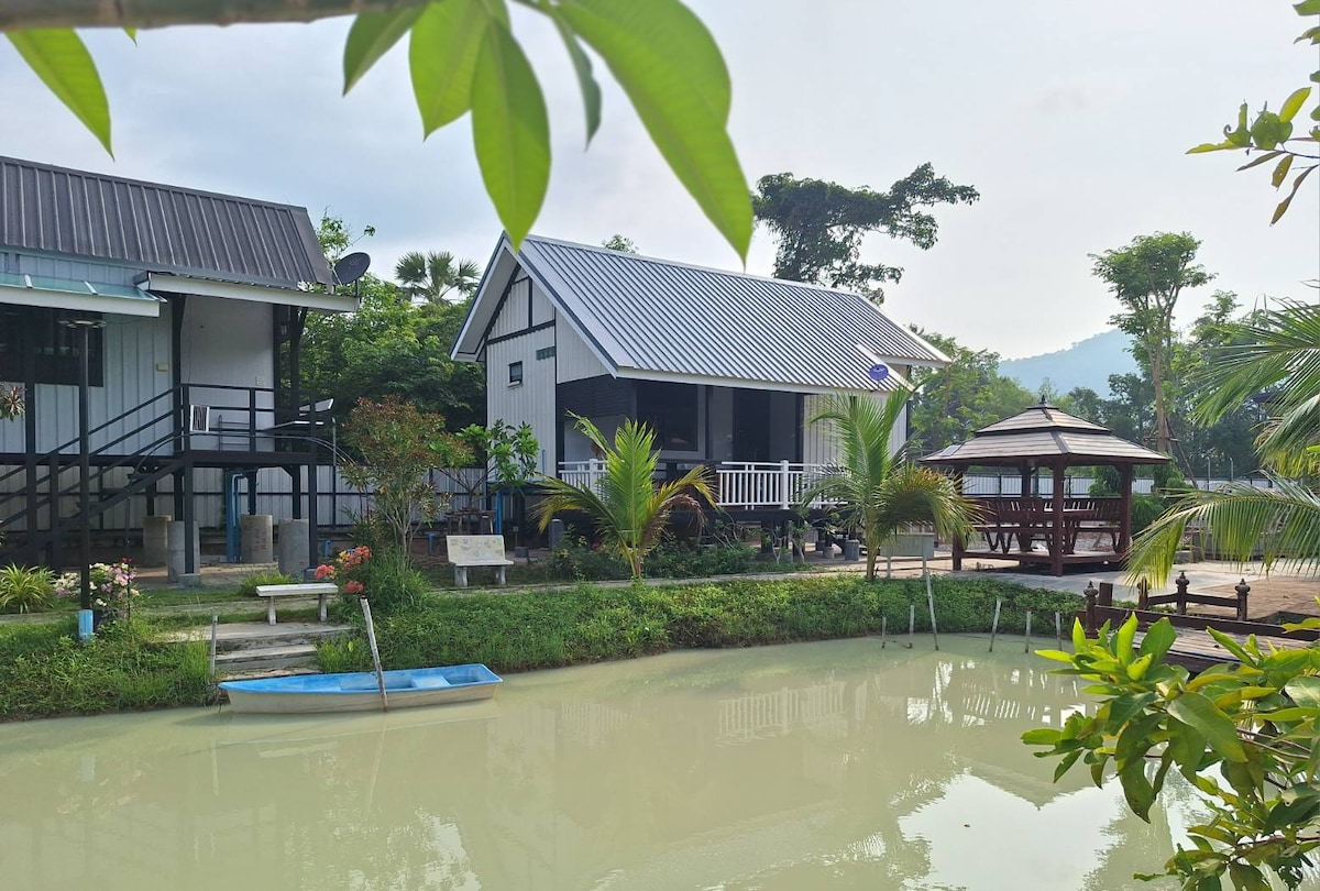 A charming homestay is nestled beside a tranquil pond, featuring a white house with a sloped roof. Surrounding greenery adds a natural touch, while a gazebo is visible nearby, complemented by a small boat resting on the water's edge.