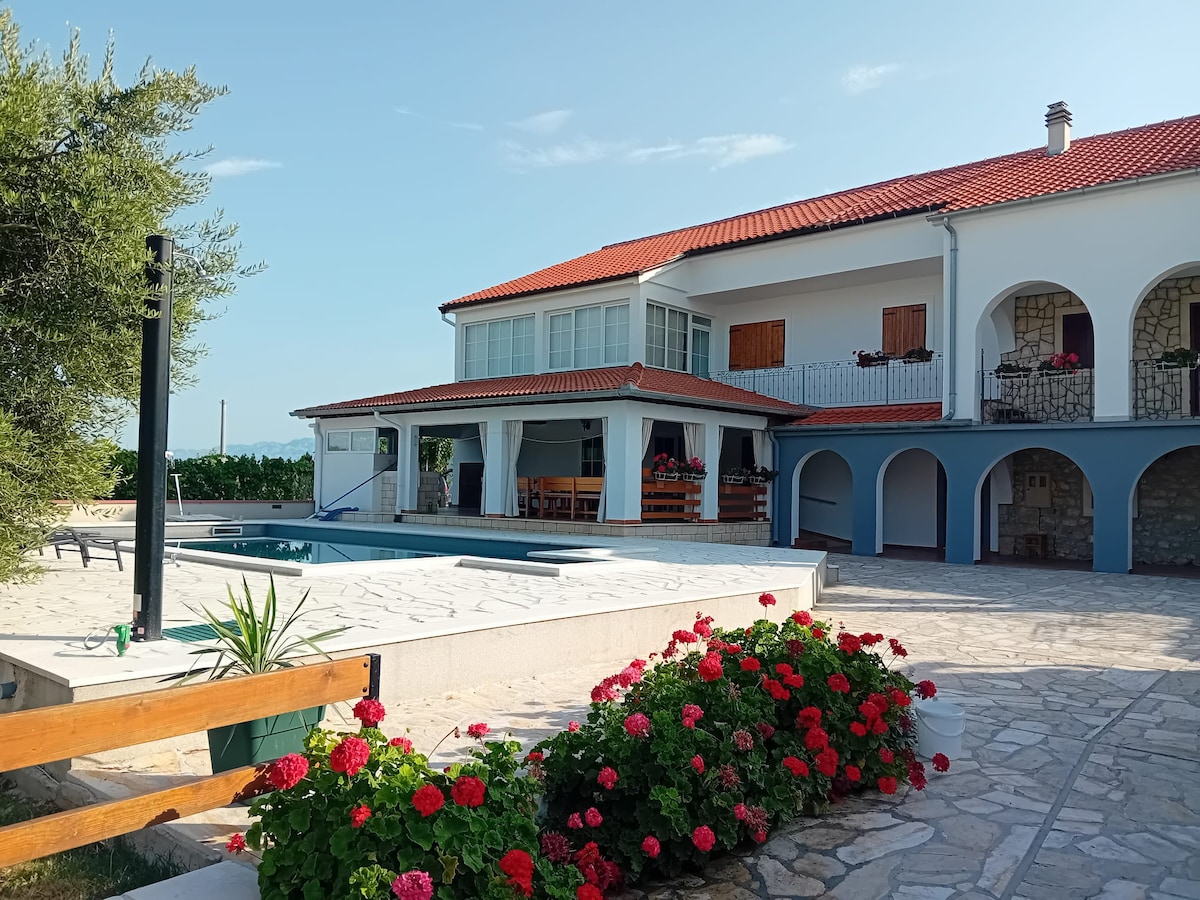 A spacious villa features a bright terrace with a private pool surrounded by well-maintained gardens. The exterior showcases a combination of stone and white stucco, with wooden shutters framing large windows. A covered seating area is positioned near the pool, offering shaded relaxation.