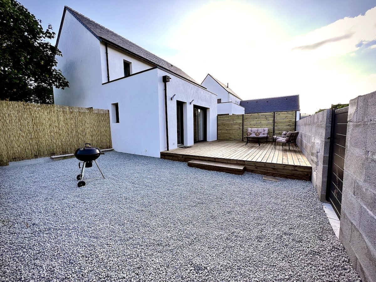 An outdoor area features a wooden deck and cozy seating, surrounded by a gravel surface. A barbecue grill is placed nearby, and natural greenery provides a backdrop. The modern white building stands in the distance, complemented by a fence for privacy.