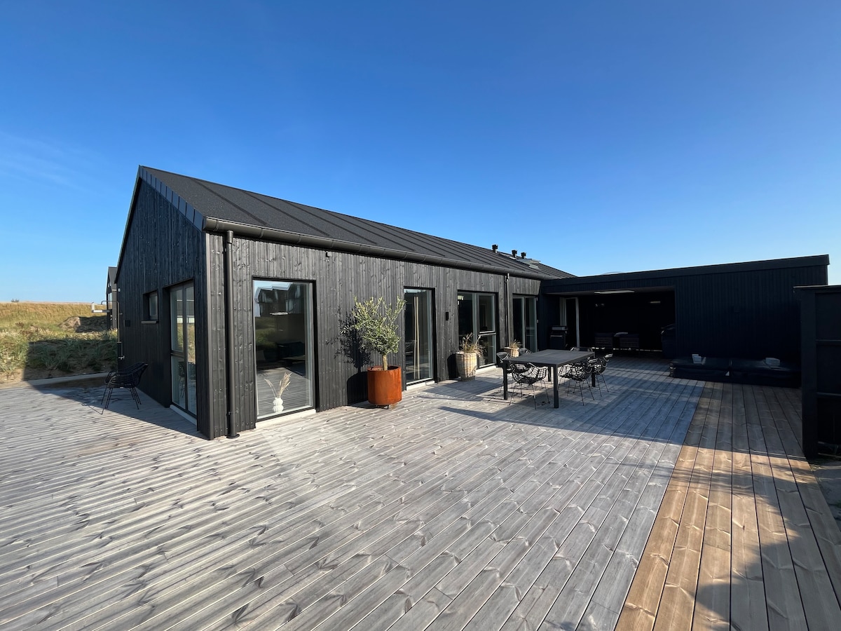 The exterior of a modern black house is shown, featuring large windows and an expansive wooden terrace. A dining table with chairs is positioned outside, surrounded by greenery. The clear blue sky complements the sleek architecture of the home.