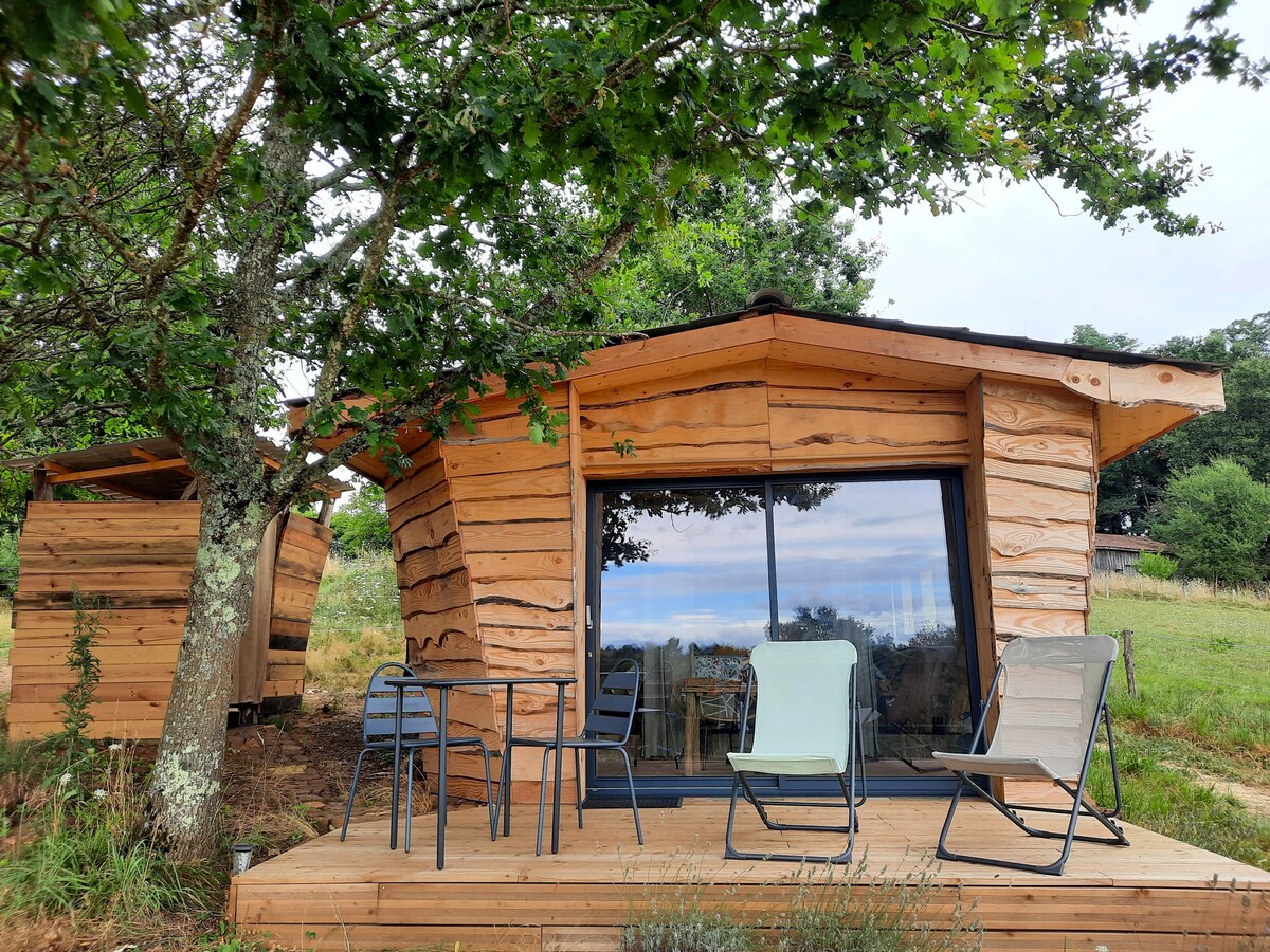 A wooden lodge with a unique design is set on a wooden deck, featuring two chairs and a small table. Large windows offer views of the surrounding greenery, allowing natural light to fill the interior space. Nearby, a second structure is visible among the trees.