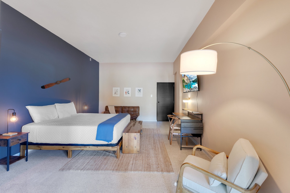 A spacious studio apartment features a large bed with crisp white bedding against a two-tone wall. A comfortable sofa is positioned nearby, with a wooden bench and a modern desk. A floor lamp illuminates the area, and a flat-screen TV is mounted on the wall.