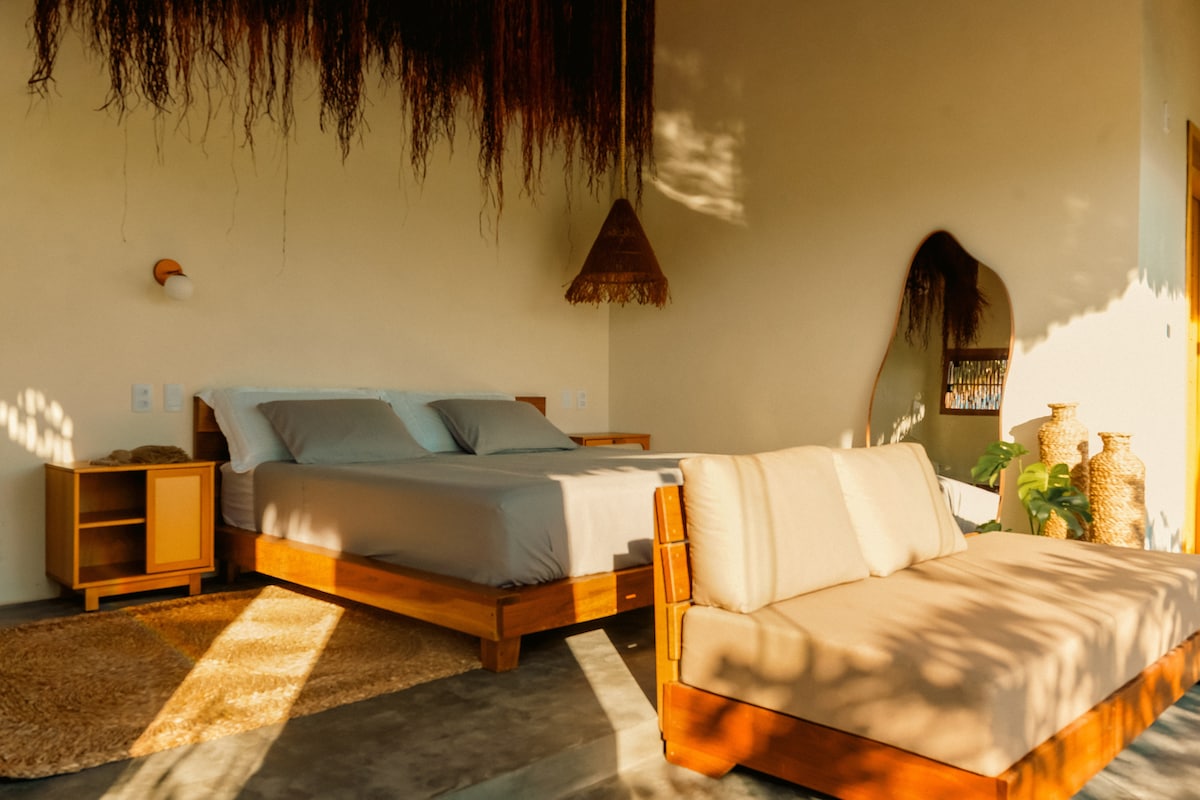 A bright sleeping area features a spacious bed with neutral bedding, complemented by a cozy seating area. Natural light streams in, creating a warm ambiance. Organic textures are present through woven decor, and a large mirror enhances the sense of openness in the room.