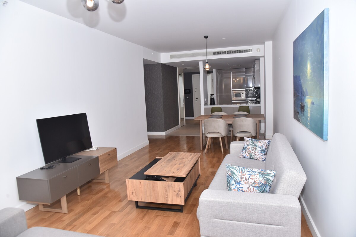 A spacious living area is presented with light wood flooring and minimalistic furniture. A comfortable sofa and armchairs are arranged around a coffee table. A flat-screen television is positioned against the wall, and a dining table is visible in the background, alongside an open kitchen.