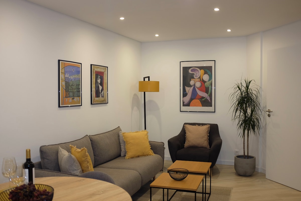 A modern living area features a gray sectional sofa with yellow accent pillows, complemented by a dark armchair. Two wooden coffee tables are arranged in front, with a decorative bowl atop. Artistic wall hangings add color, and a potted plant introduces a touch of nature.