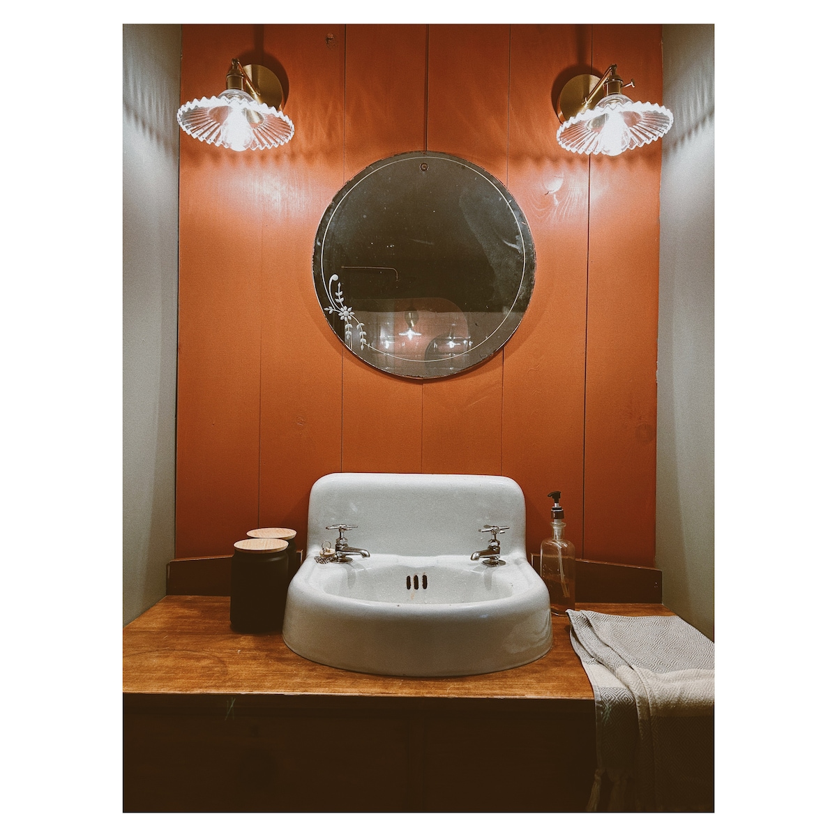 A bathroom space features a vintage-style sink with dual faucets, positioned below a circular mirror. The walls are painted in a warm orange hue, and two light fixtures provide a soft glow, adding to the inviting ambiance. Homemade wooden cabinetry completes the design.