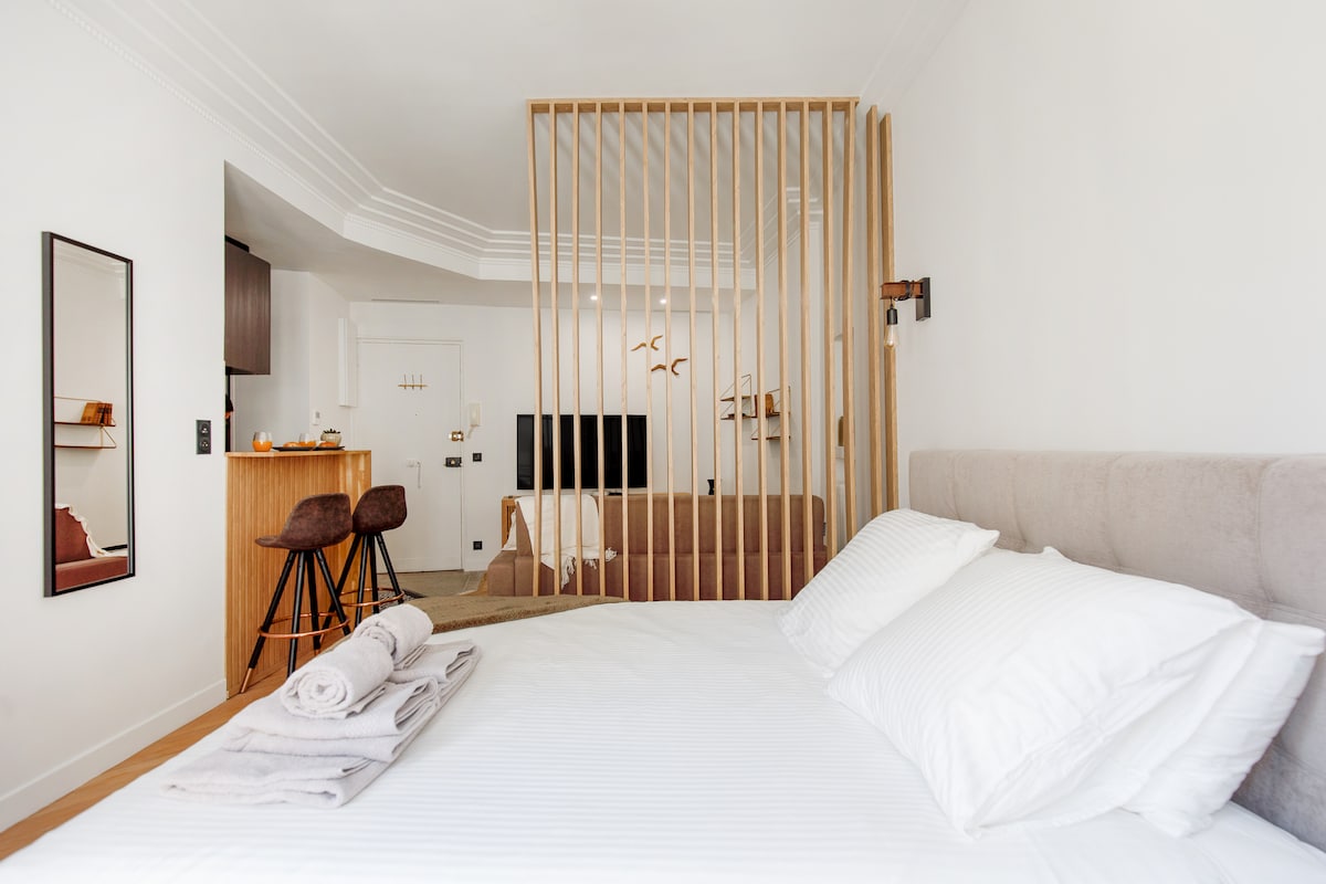 A well-lit apartment showcases a spacious bed with crisp white linens. A wooden partition separates the sleeping area from an inviting living space that includes a bar with stools. Decorative elements and a mirror enhance the minimalist design, creating an open atmosphere.