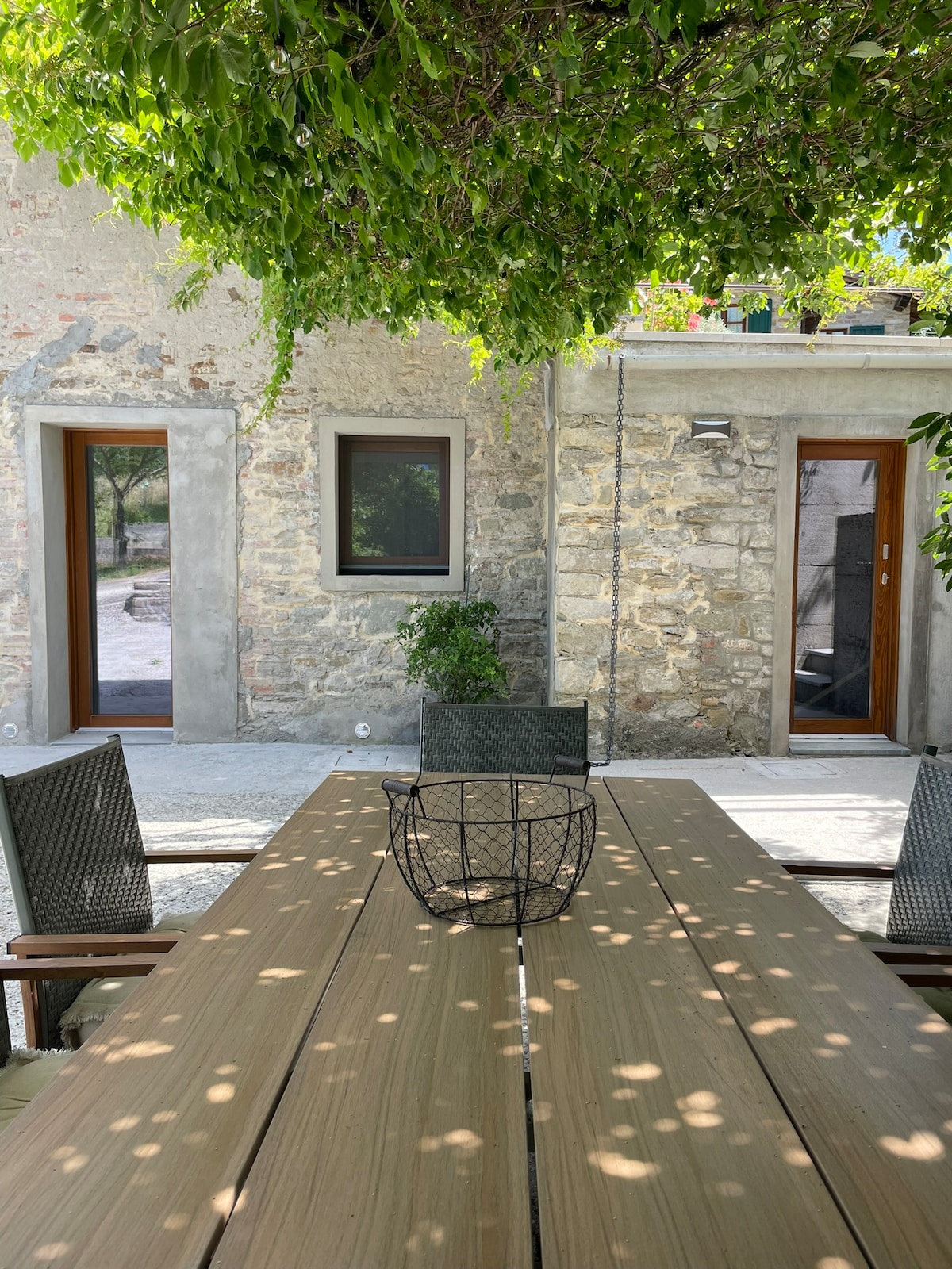 A wooden table is positioned outdoors under lush greenery, with an arrangement of chairs around it. Stone walls of the rustic building provide a natural backdrop, and two doorways lead to private access points.