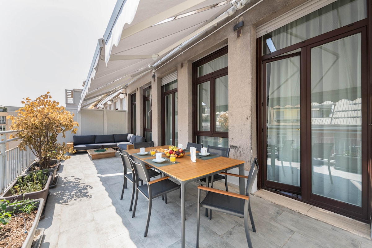 A spacious terrace features a large wooden dining table surrounded by six grey chairs. A comfortable seating area with dark cushions is positioned against a wall. Potted plants and a low stone wall provide a natural touch, while the canopy overhead offers shade.