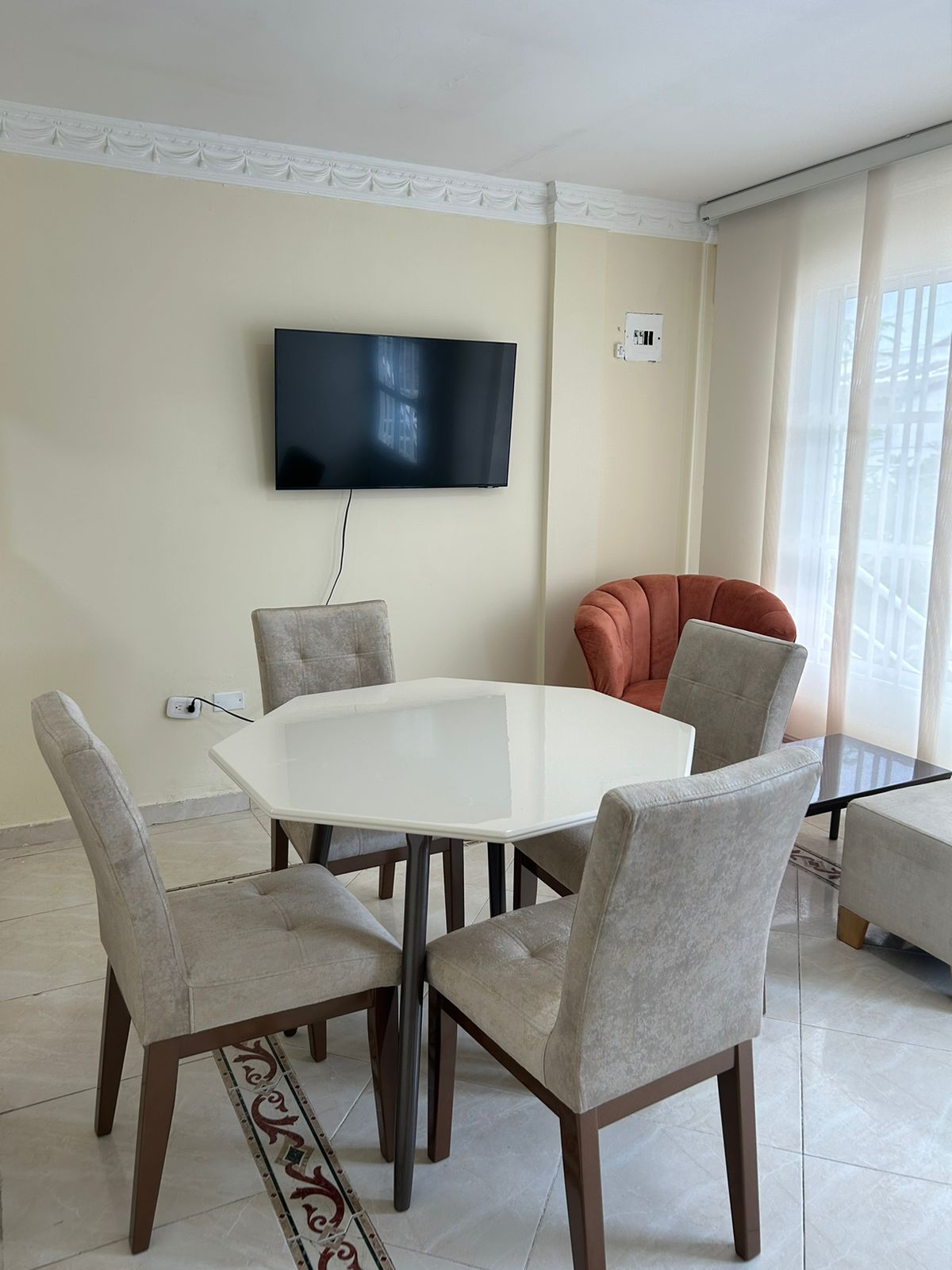 A dining area features a white hexagonal table surrounded by four upholstered chairs. A sleek television is mounted on the wall, and natural light filters through the sheer curtains, illuminating the space. A cozy orange armchair is positioned nearby for additional seating.