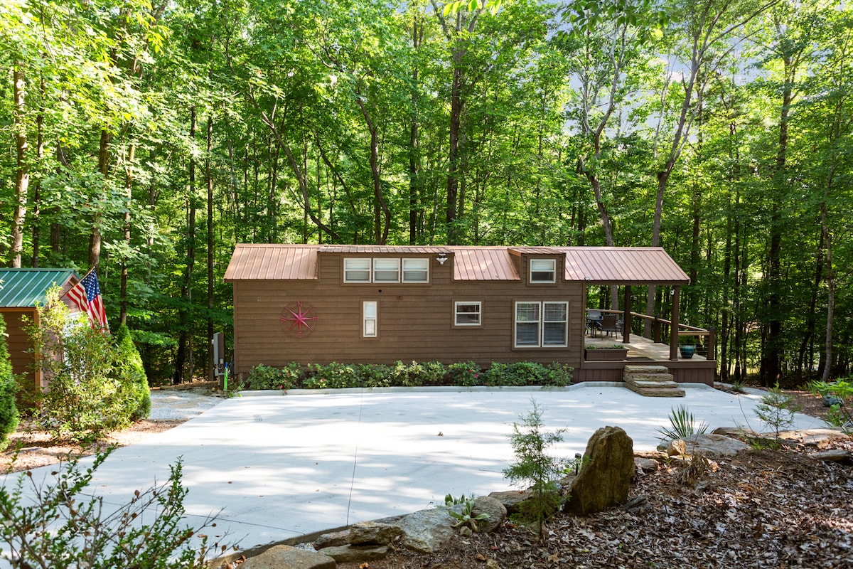 The cabin is set amidst a lush wooded landscape, featuring a sturdy exterior with a warm brown finish. It includes a metal roof and large windows, offering natural light. A spacious driveway leads to the entrance, providing convenient access to the inviting outdoor deck.