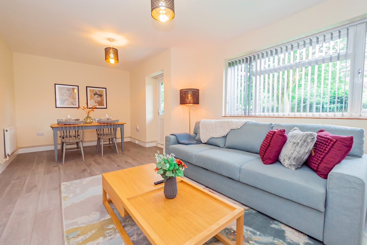 A spacious living area is featured, furnished with a light blue sofa and a wooden coffee table. A dining area with a table and chairs is visible in the background. Natural light filters through large windows with vertical blinds, complementing the neutral walls.