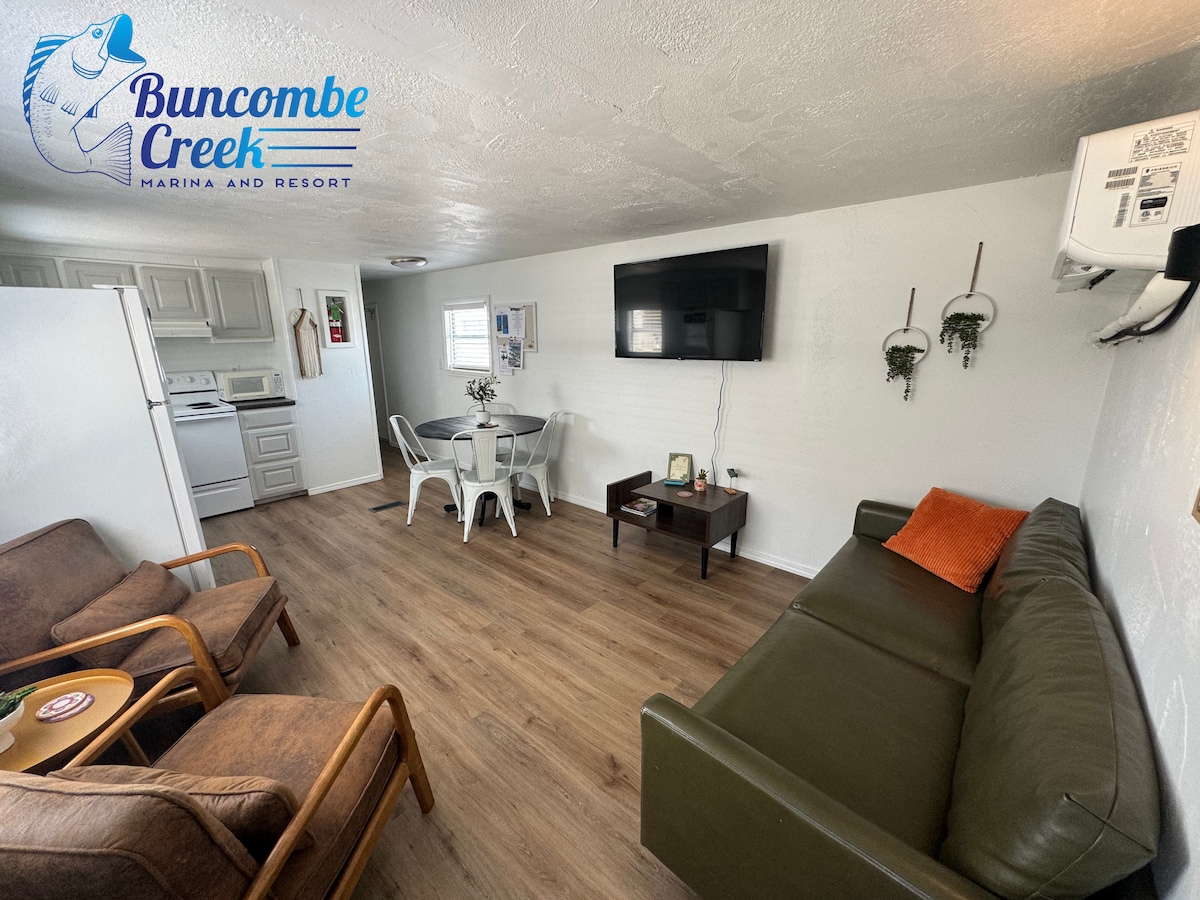 A cozy living area features a green sofa and two brown chairs, arranged around a wooden coffee table. A dining table with white chairs is positioned nearby. A wall-mounted television is present, and natural light enters through the window. The flooring is a warm wood tone.