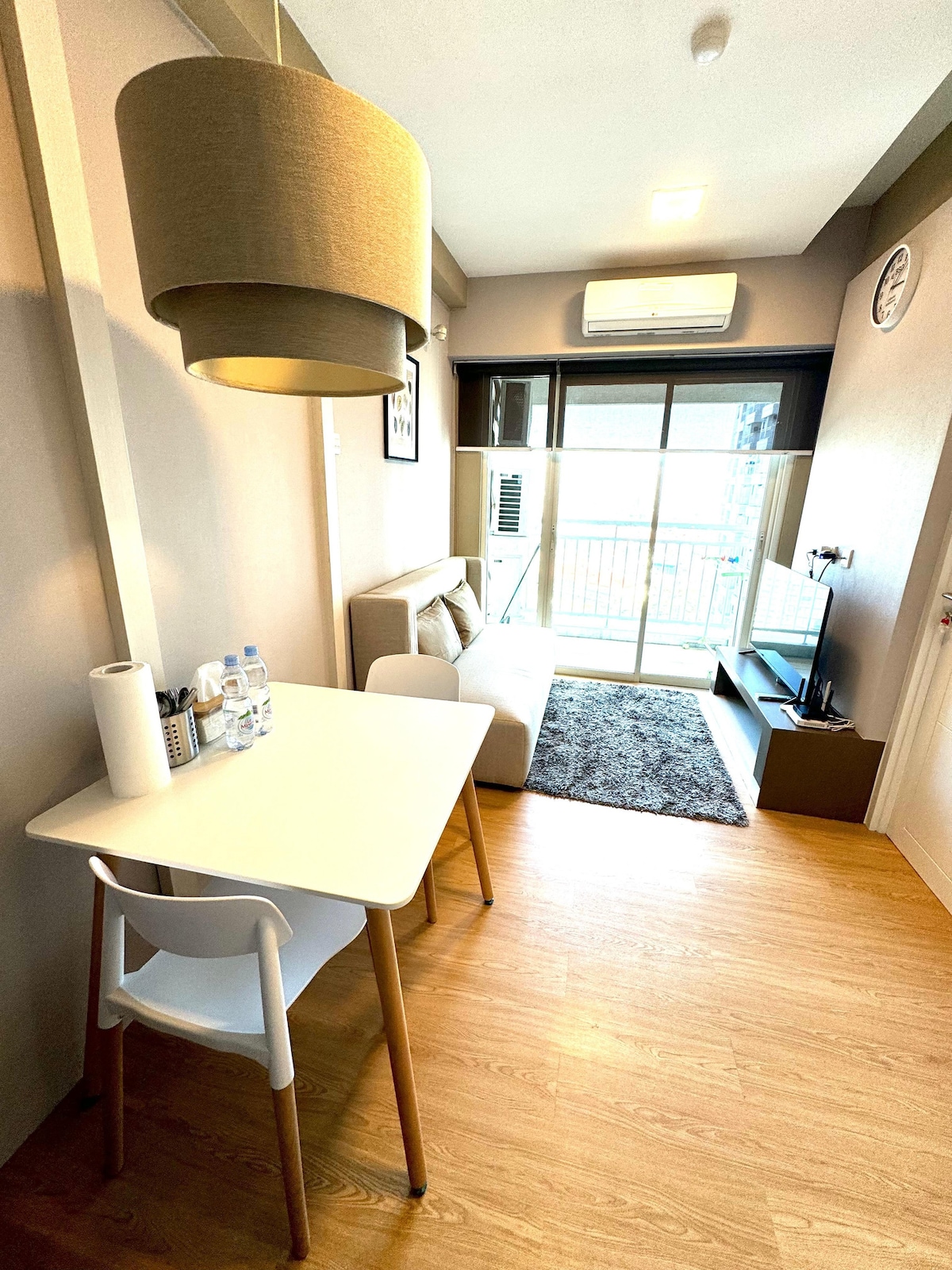 A compact living space features a small white table with wooden legs accompanied by a white chair. A beige sofa is positioned near a large window, offering access to natural light. The floor is covered with light wooden flooring, while a dark rug adds contrast.