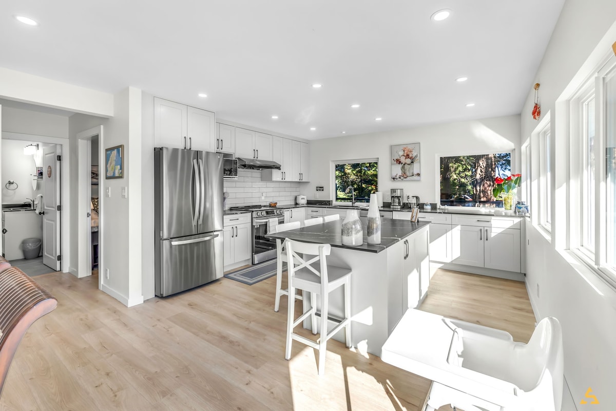 An open-concept kitchen features sleek white cabinetry and stainless steel appliances. A central island provides ample counter space and seating. Natural light fills the room through large windows, illuminating the modern design with a minimalist style and wooden flooring.