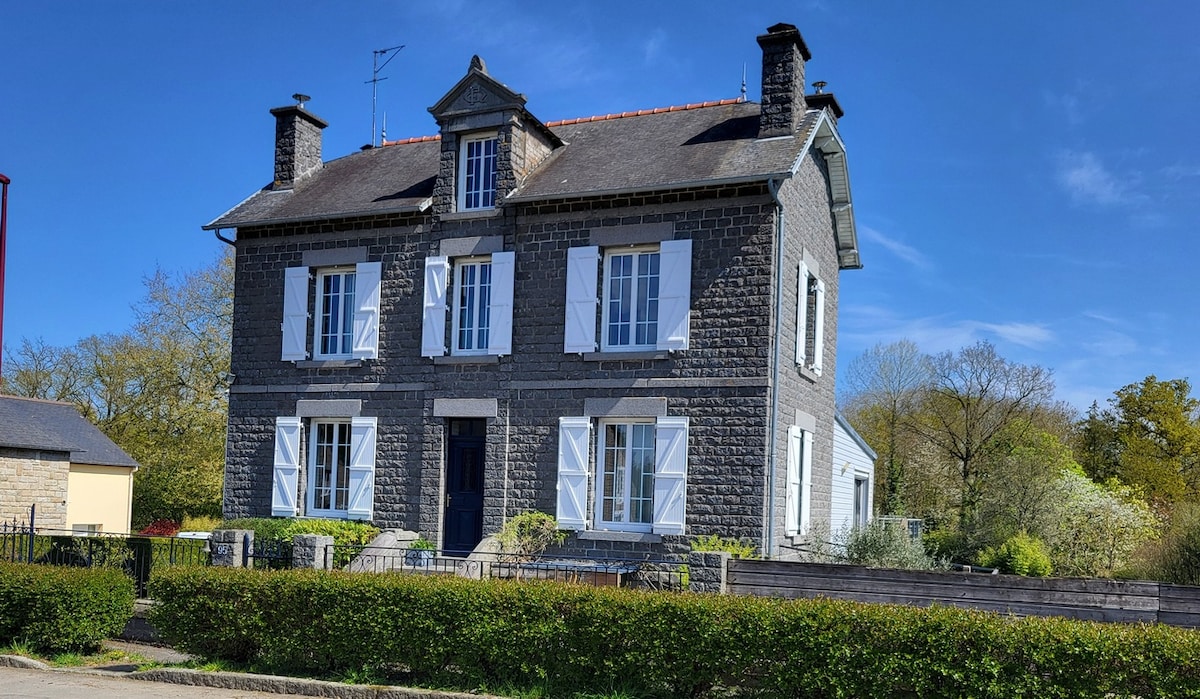 The two-story family home displays a classic stone facade, featuring white shutters and a central blue door. Surrounding the property, a neatly trimmed hedge frames the entrance, while trees and greenery provide a natural backdrop against a clear blue sky.