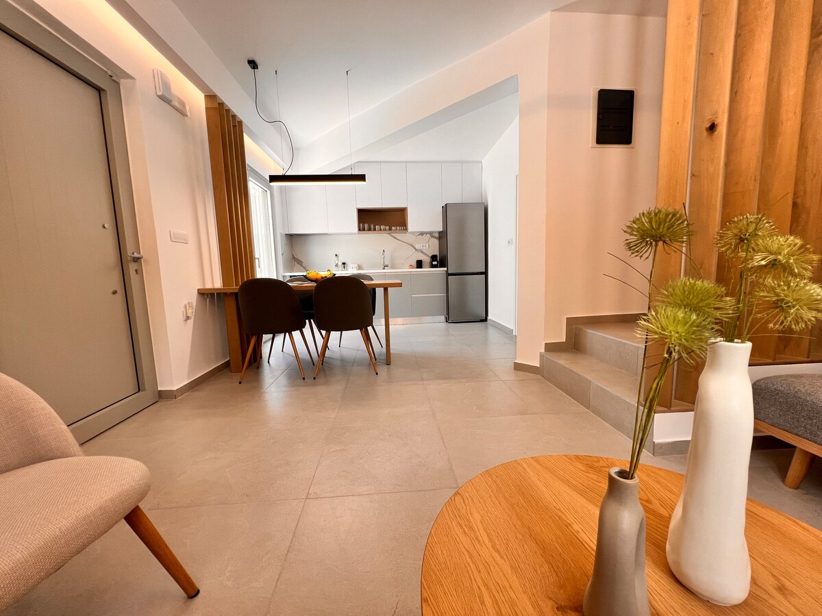 A contemporary living space is presented with a modern kitchen and dining area. A wooden dining table is surrounded by four chairs, with a view of a well-equipped kitchen. Soft lighting complements neutral tones in the decor, enhancing a serene ambiance.