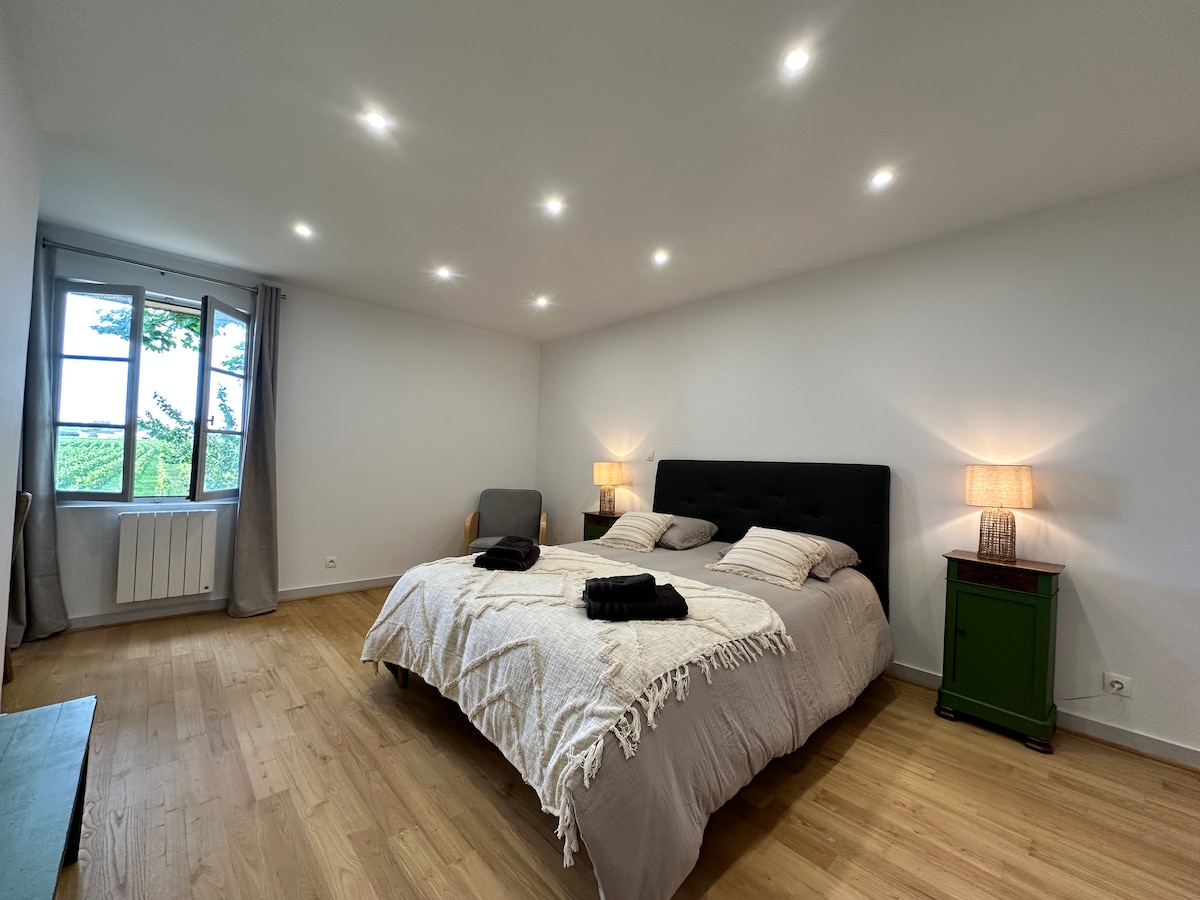 The spacious bedroom features a large bed with neutral linens and two bedside lamps providing soft lighting. A comfortable armchair is positioned nearby, and a window offers a view of the surrounding greenery.