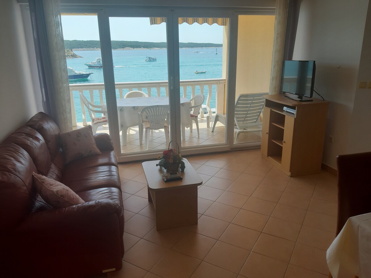 A living area features a maroon leather sofa and a wooden coffee table, positioned near expansive glass doors that open to a balcony with views of the water. A flat-screen TV is placed on a light wood cabinet, complementing the neutral tile flooring.