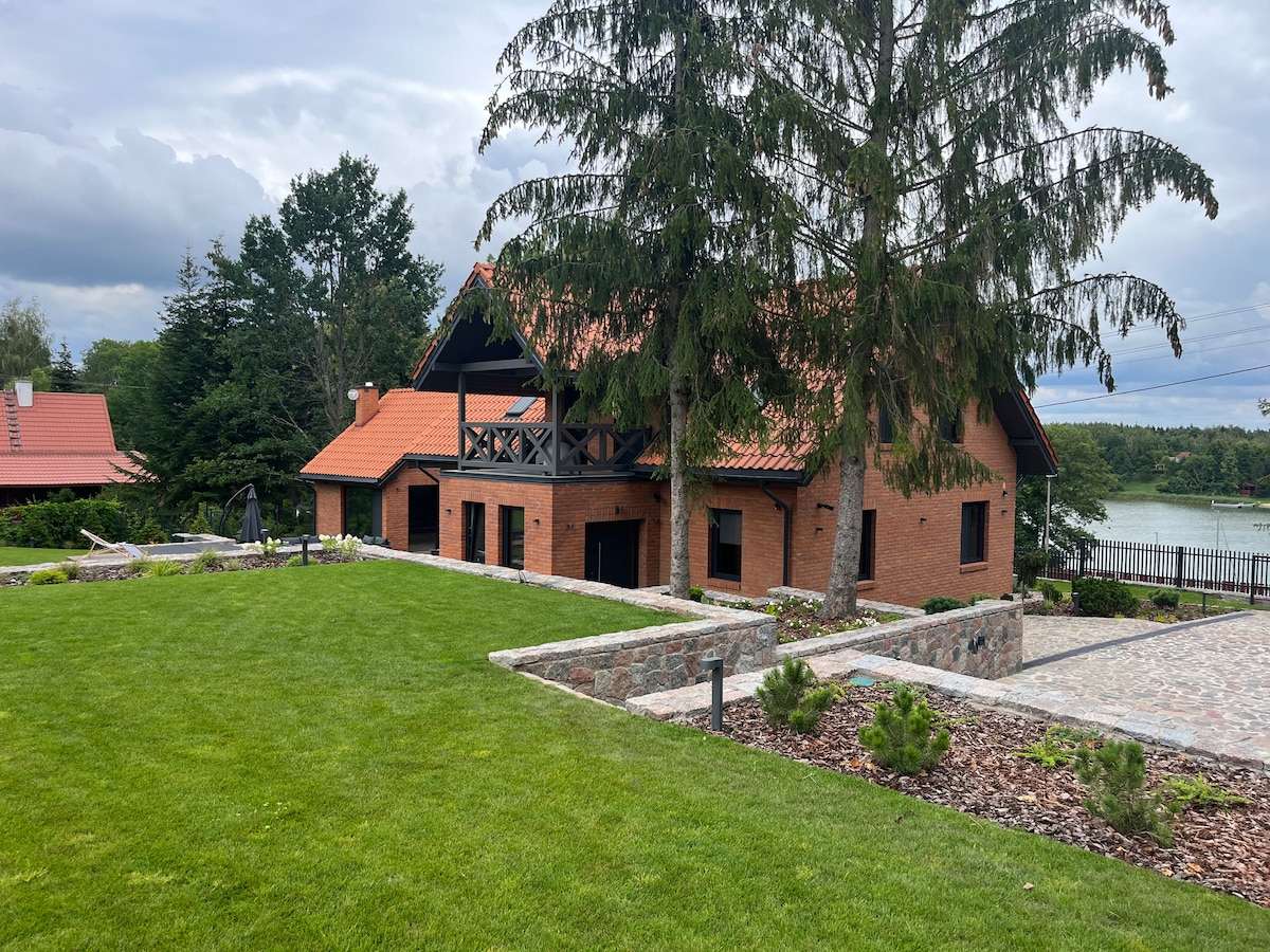 A spacious outdoor area is showcased, featuring a well-maintained lawn with various landscaping elements. A rustic brick building with a sloped roof and large balcony is visible. Surrounding trees provide shade, while a view of the water can be seen in the background.