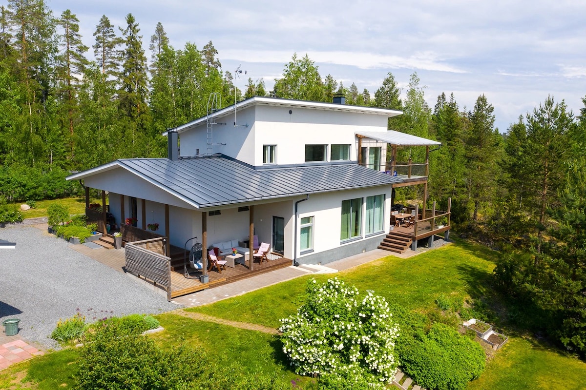 A modern two-story house is positioned within a lush, green landscape. The exterior features a blend of wood and large glass windows. A spacious deck extends from the house, surrounded by trees, with outdoor seating visible on the patio.