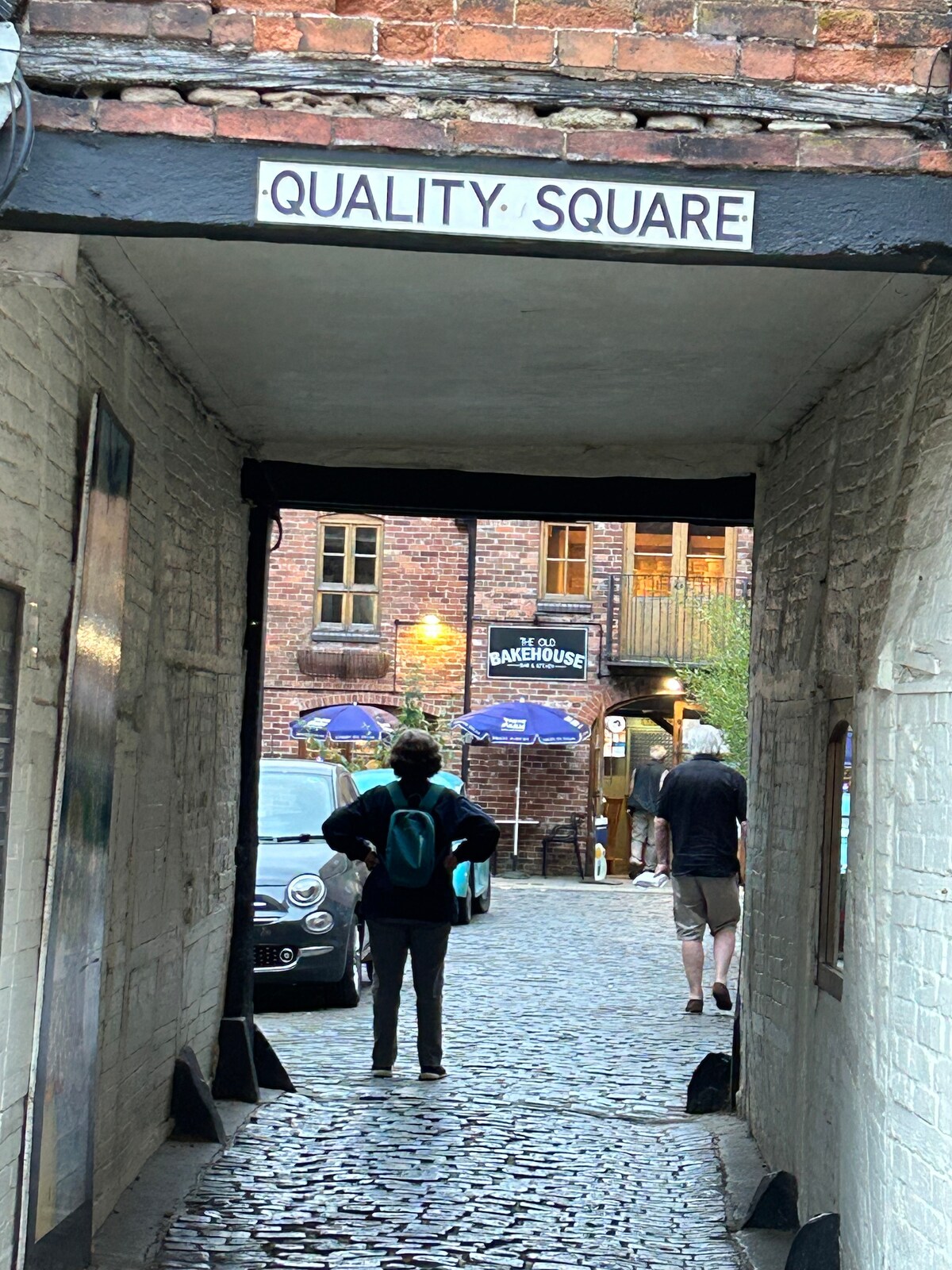 An archway labeled 'QUALITY SQUARE' leads into a cobbled courtyard. Two individuals are visible, one standing with a bag, while another walks away. The background features a building with outdoor seating under umbrellas and a parked car.