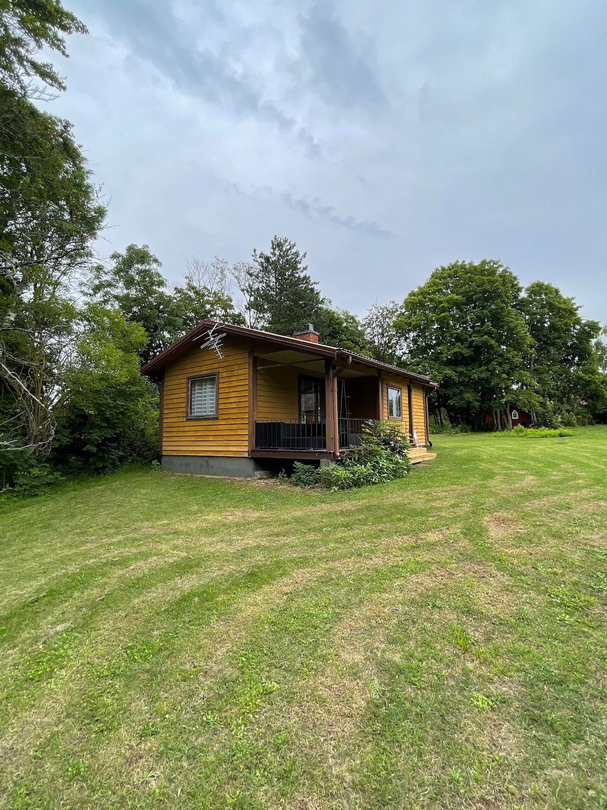 A charming yellow cottage is nestled among lush greenery, featuring a welcoming porch area. Surrounded by a spacious lawn, the structure is positioned to invite relaxation in a natural setting. Trees and shrubbery add to the serene ambiance of this peaceful getaway.