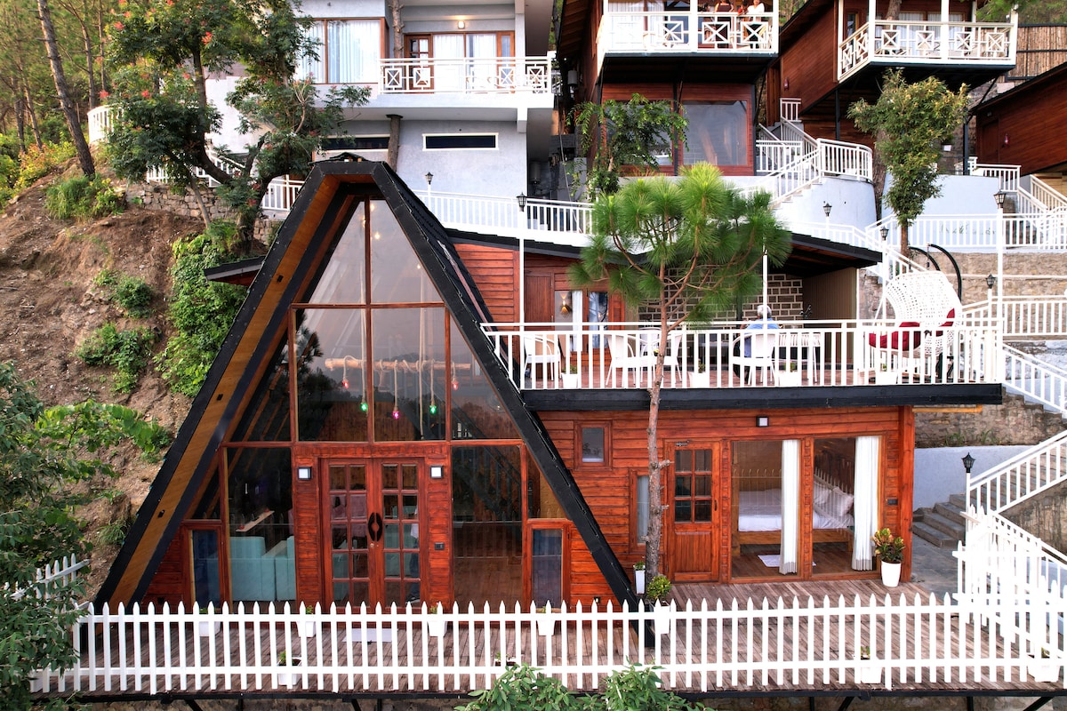 The A-frame wooden villa features a sloping roofline and expansive glass windows, allowing natural light to flood the interior. A white picket fence surrounds the property, providing a charming entrance. Green trees and a rocky hillside create a natural backdrop, enhancing the secluded ambiance.