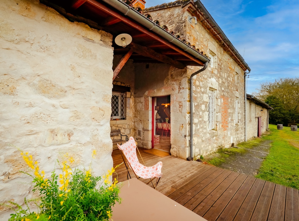 An exterior view of a rustic stone house is showcased, featuring a wooden deck with a single patterned chair. A flower pot filled with yellow blooms sits prominently, and the lush green lawn extends beyond the structure under a partly cloudy sky.