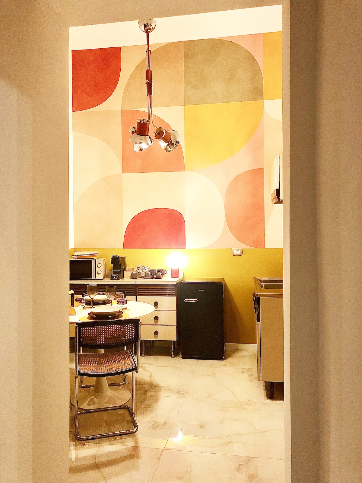 A bright kitchen area is visible through the doorway, featuring a geometric wall design in warm tones of red, yellow, and beige. A small dining table with metallic chairs is set beside modern appliances, including a microwave and a compact refrigerator.