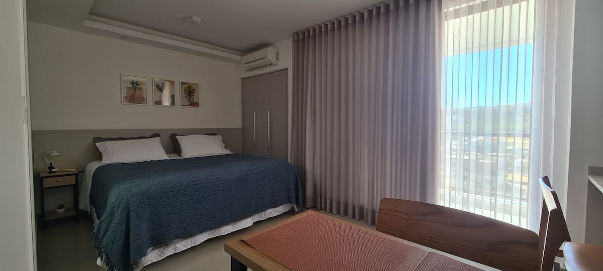 The well-lit studio features a spacious bed layered with a navy blue coverlet and white pillows. A desk is positioned nearby, and large windows with sheer curtains allow natural light to fill the room, offering a view of the outdoor surroundings.