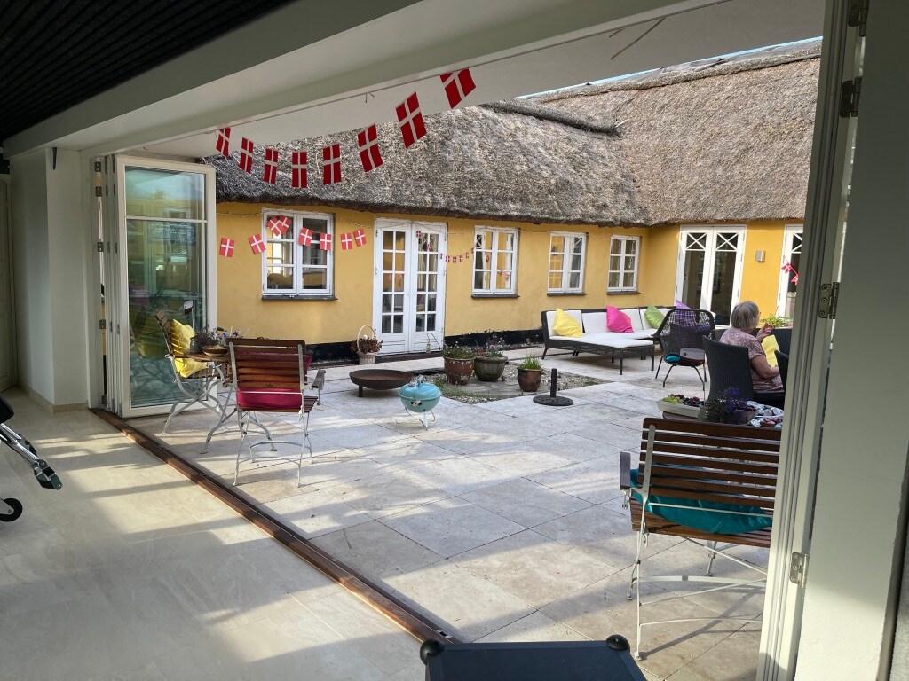 A charming outdoor terrace is framed by spacious glass doors, creating a seamless transition from indoors. Colorful seating options are scattered throughout, complemented by potted plants. A thatched-roof building with warm yellow walls provides a welcoming backdrop to the scene.