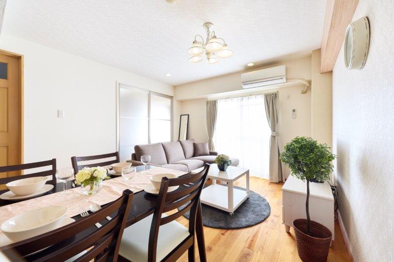 A bright living area is depicted with a light-colored sofa and a simple coffee table on a circular rug. A dining table is set for six, showcasing plates and glasses. Natural light enters through large windows with sheer curtains, enhancing the welcoming environment.