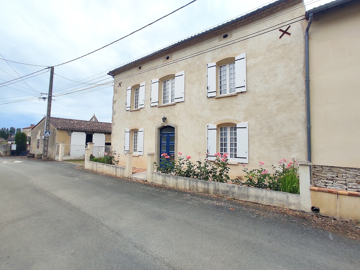 The exterior of a charming country house is depicted, featuring a pale stucco facade with white shutters. A vibrant blue door serves as the focal point, flanked by flower beds filled with pink blooms. The surrounding area is peaceful, with a clear view of the road and nearby structures.